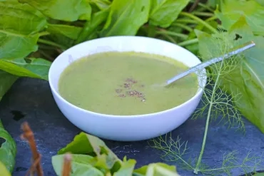 Une assiette creuse contenant de la soupe verte et une cuillere est installée sur une ardoise avec des légumes verts autour