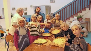 deux adultes et un groupe d'enfants avec charlottes et tabliers montrent leurs pizza finies en cuisine
